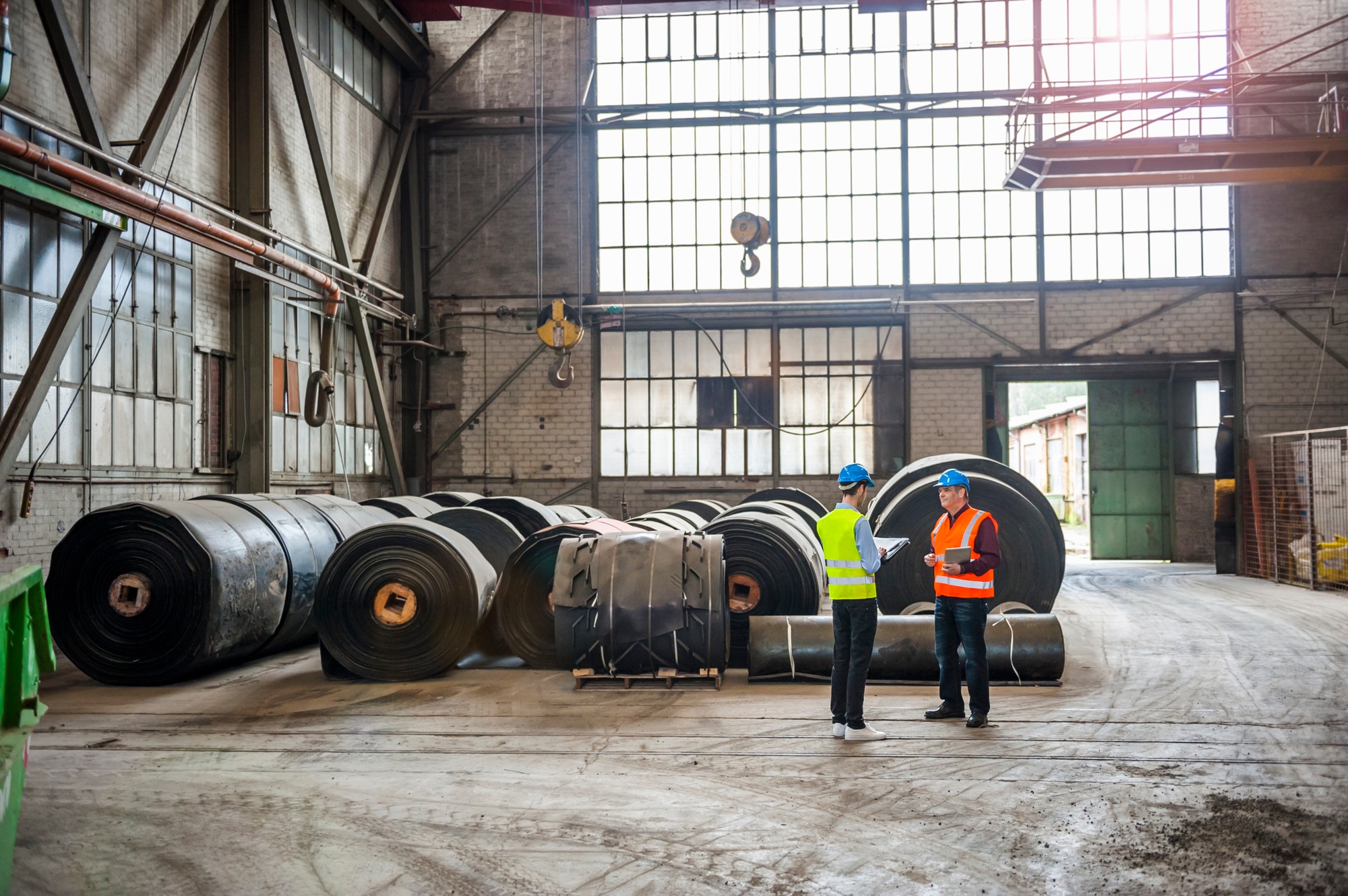 Two men with documents in factory hall with rolls of rubber